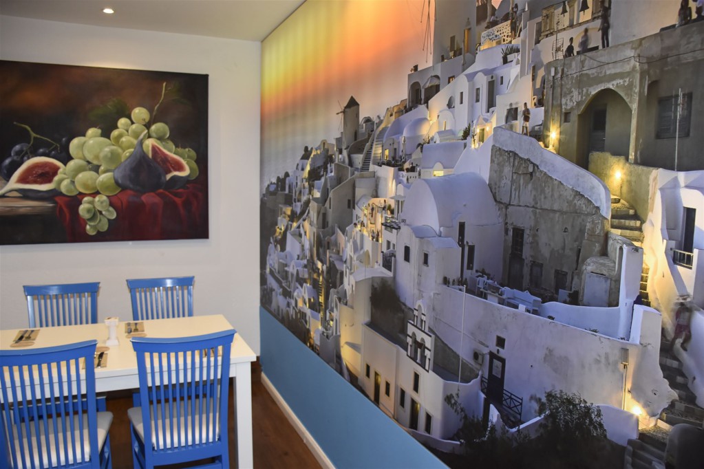 Ein Wohnraum, der an der linken Wand ein Gemälde von Trauben und Feigen zeigt und an der rechten Seite eine große Wandmalerei eines mediterranen Dorfe